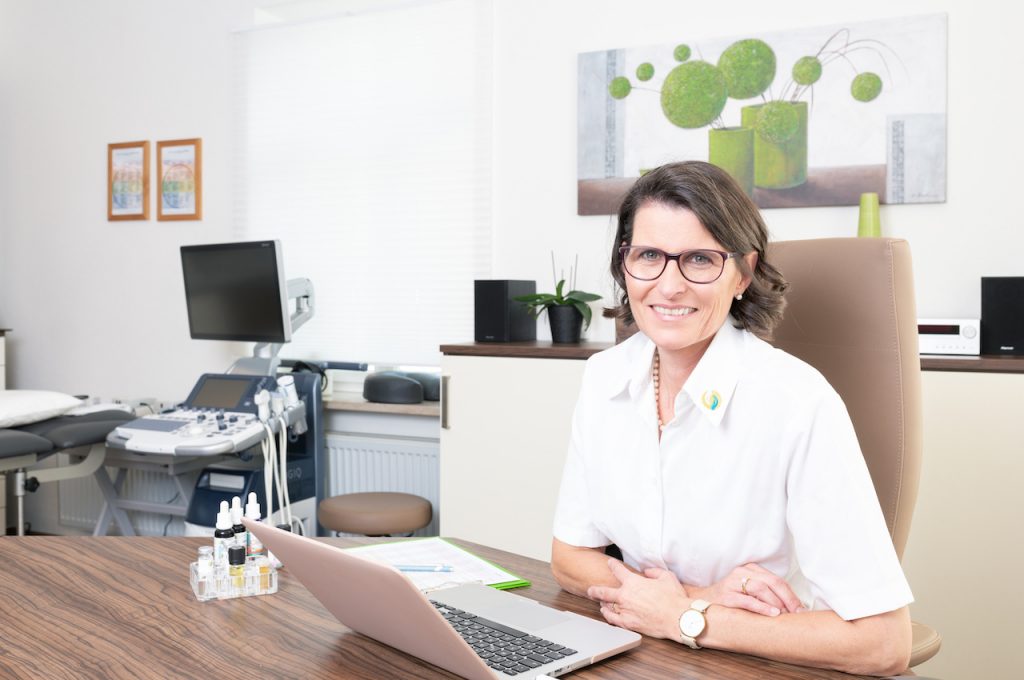 Ariane Zappe in ihrer Praxis am Schreibtisch mit Laptop vor sich und Praxisausrüstung im Hintergrund - Querformat
