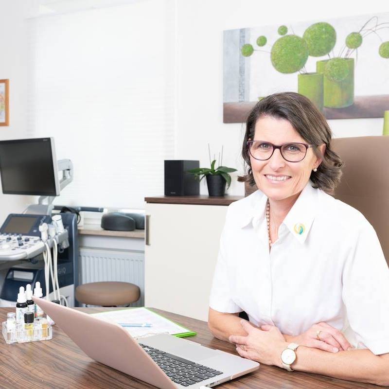 Ariane Zappe in ihrer Praxis am Schreibtisch mit Laptop vor sich und Praxisausrüstung im Hintergrund - Quadratisch